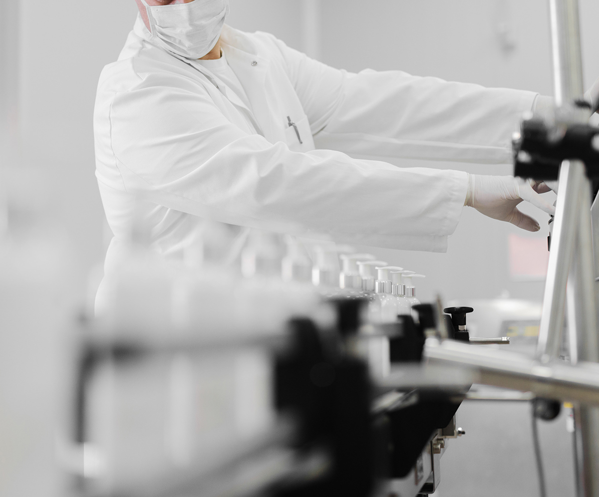 Operator in lab coat, mask and gloves adjusting a pump bottle filling line